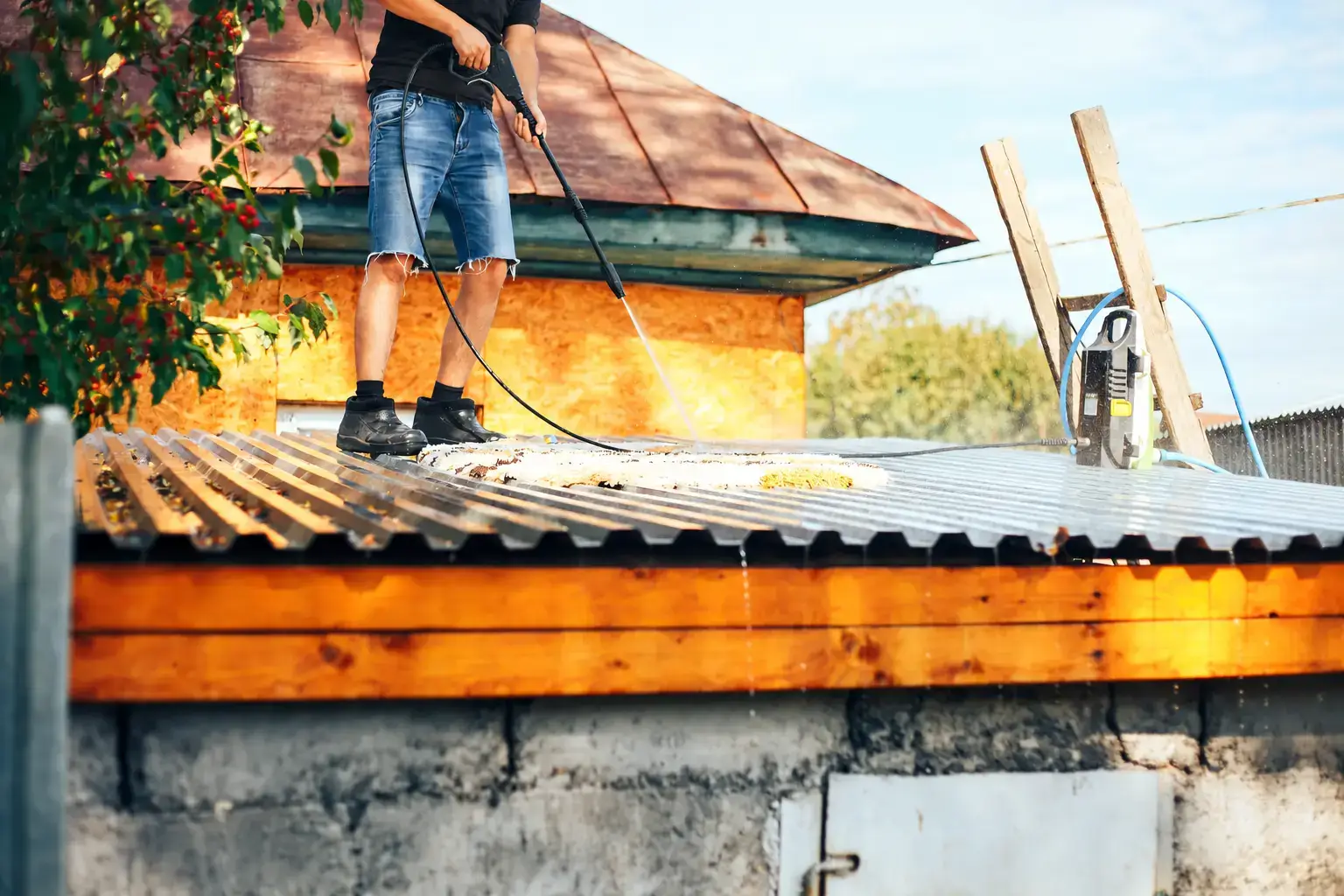 Worker using a pressure washer on a corrugated metal roof.