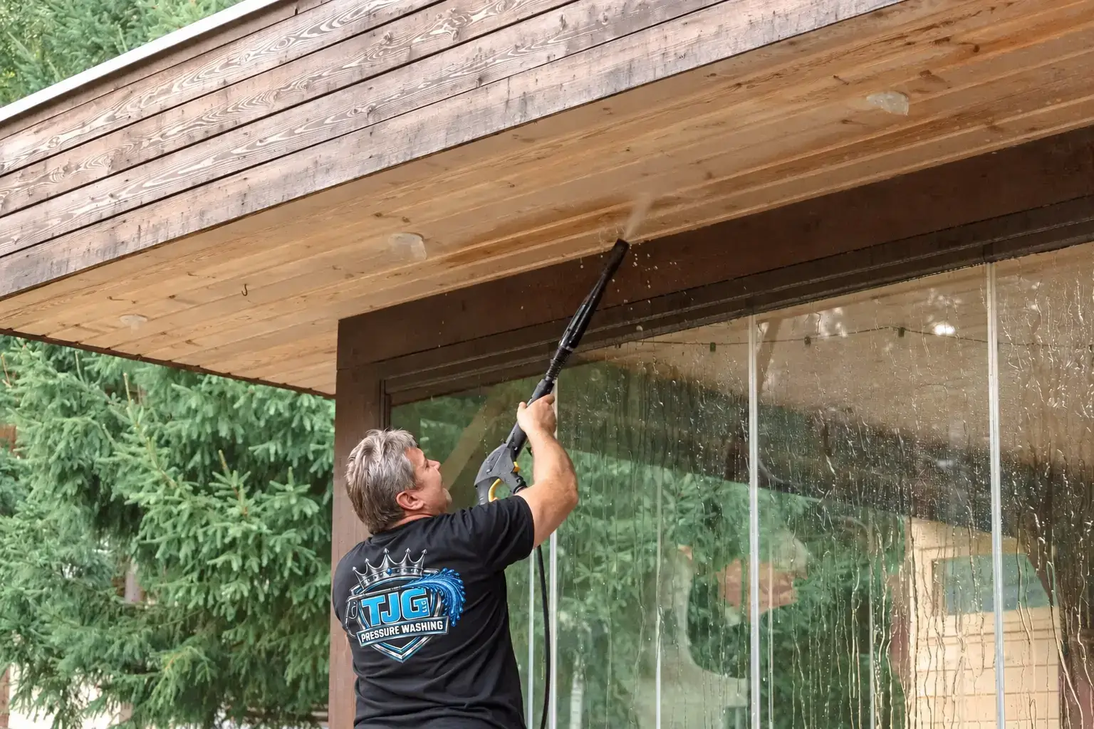 Worker in a black TJG shirt pressure washing wooden soffits.